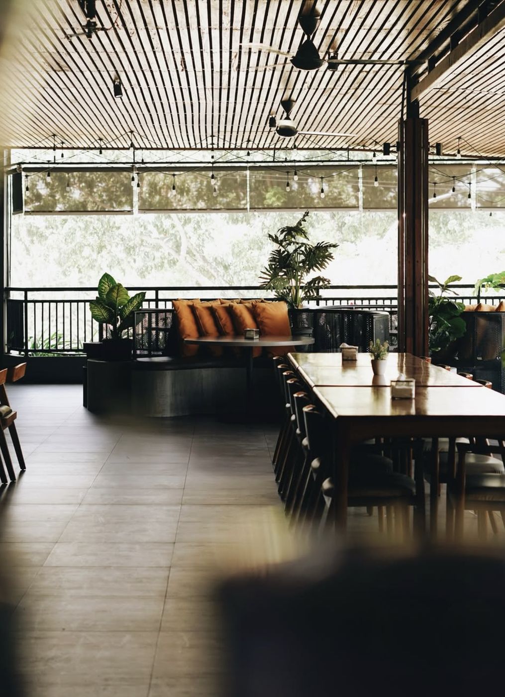 Teras semi-outdoor dengan plafon kayu, meja panjang, dan tanaman